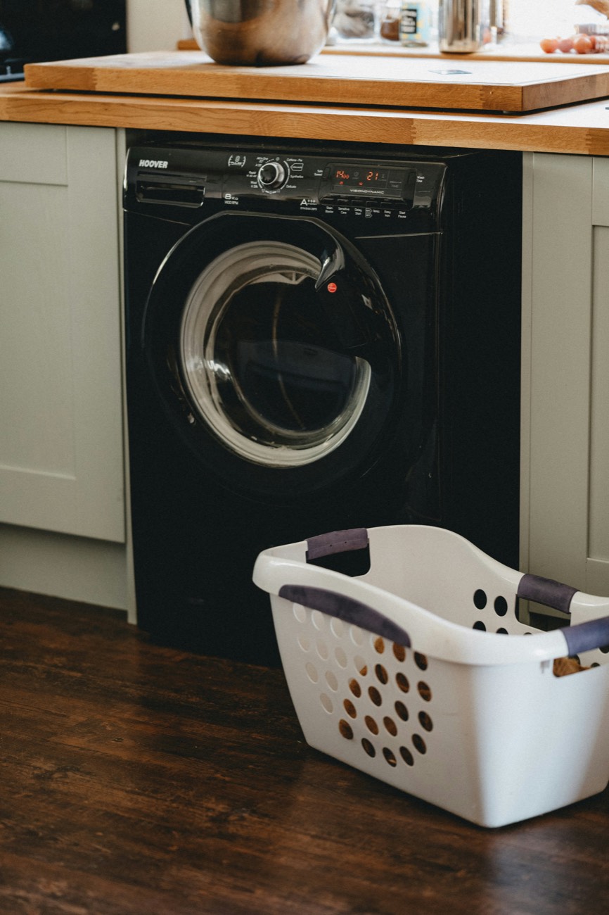 Black front-load washing machine installed under a kitchen counter, with a laundry basket in front